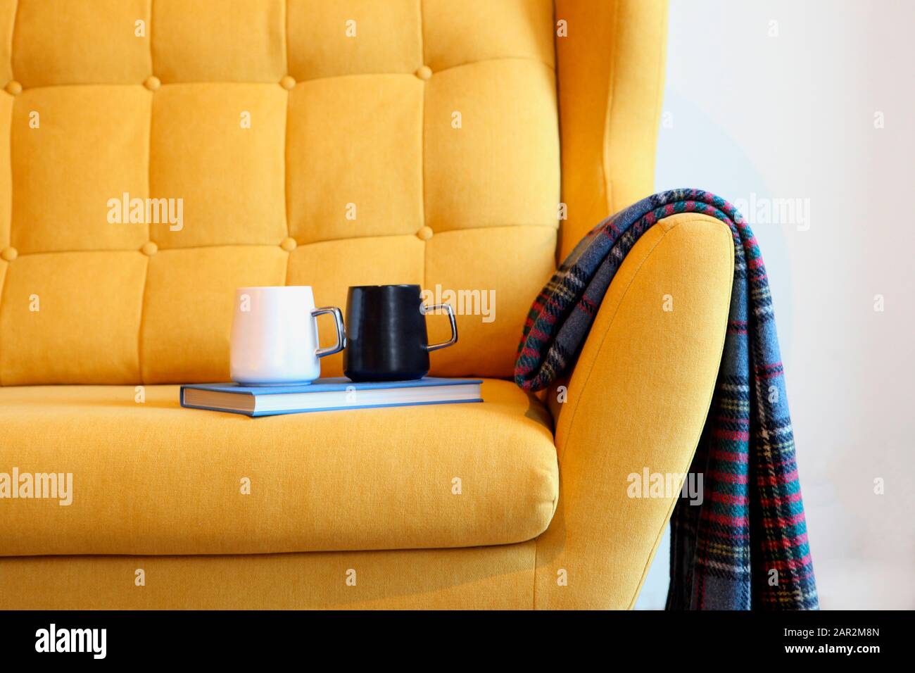 Tasse Tee und blaues Buch auf einem gelben Trainer mit Decke. Details zum Stillleben im Inneren des Wohnzimmers. Gemütliches Innenleben, Hauskomfort Stockfoto