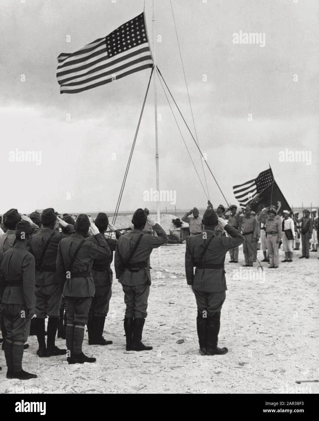 Kapitulation des Wake Atolls, 4. September 1945: Die US-Flagge über Wake Island anheben am 4. September 1945, da ein US Marine Corps Bugler "Farben" spielt. Dies war das erste Mal, dass die Stars and Stripes Wake seit ihrer Einnahme durch die Japaner am 23. Dezember 1941 überflogen hatten. Der Offizier, der im rechten Vordergrund steht, ist Rear Admiral Shigematsu Sakaibara, japanischer Befehlshaber auf der Wake. Zu den Farben, die von der US-Partei getragen werden, gehört der rechte Hintergrund, die Flagge des US Marine Corps. Stockfoto