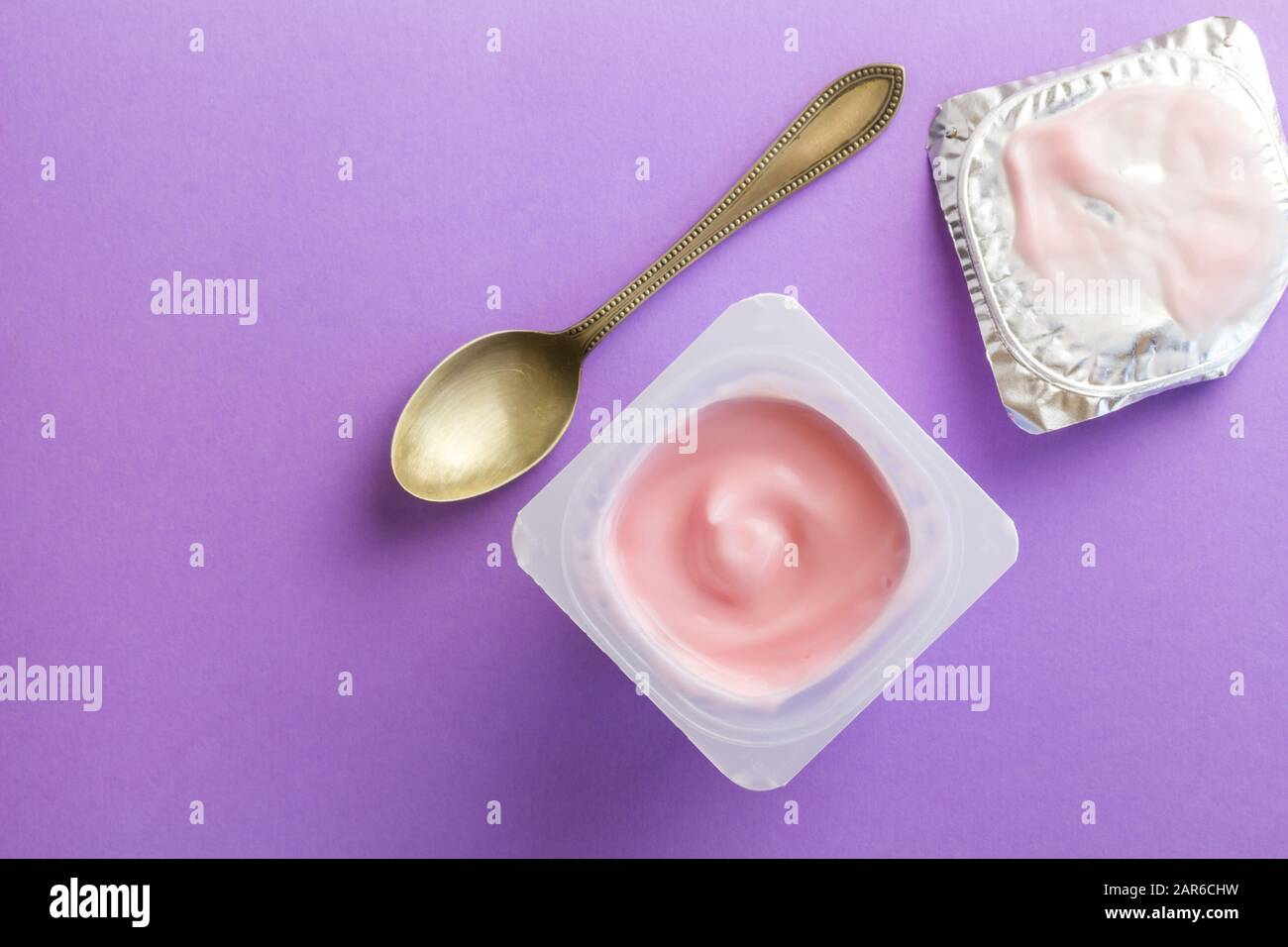 Gesunder Erdbeerobst aromatisierter Joghurt mit natürlicher Farbgebung in Kunststoffbecher isoliert auf violettem Hintergrund mit kleinem Löffel und Foliendeckel - Draufsicht Stockfoto