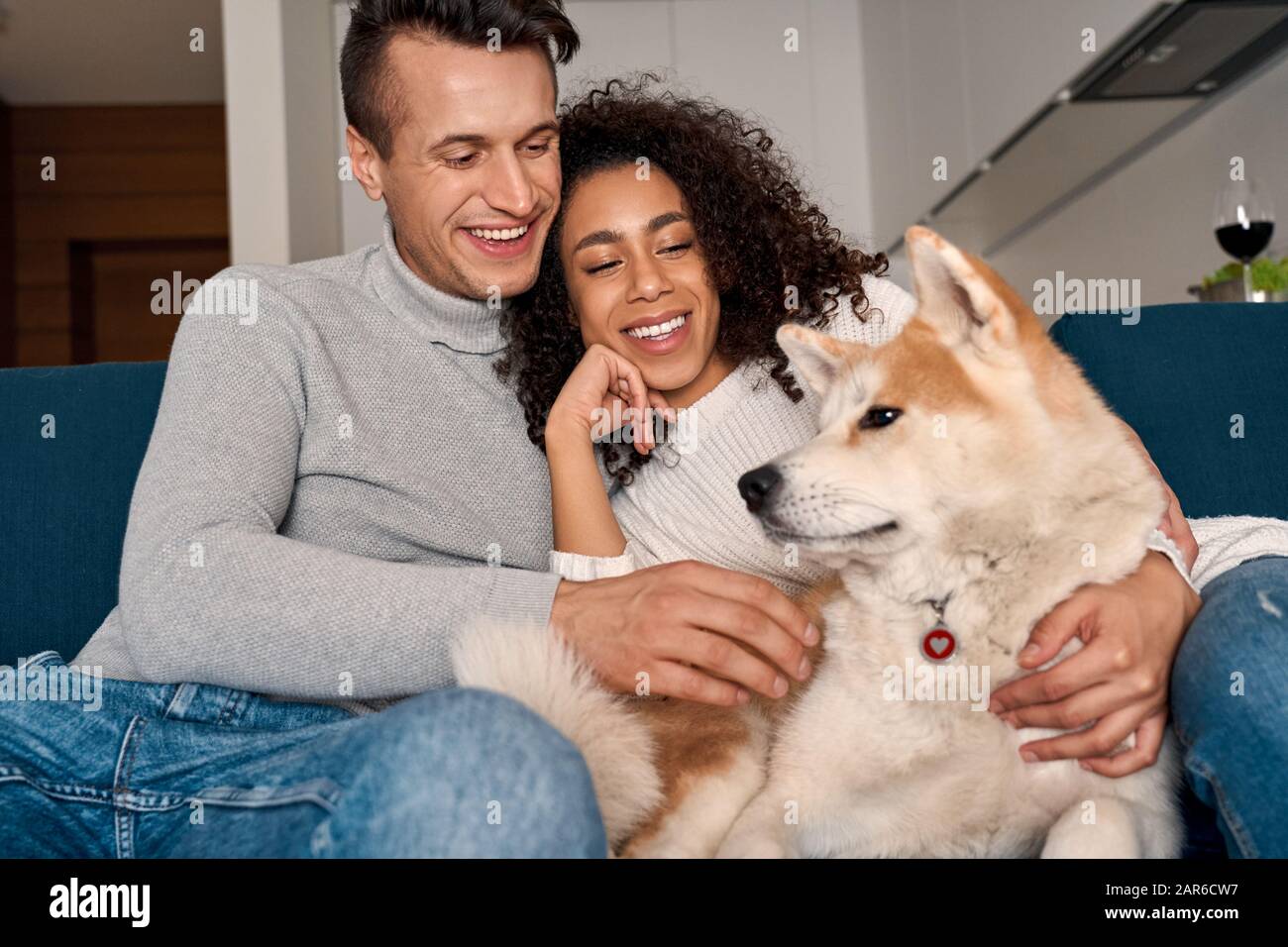 Romantisches Datum. Junges multiethnisches Paar zu Hause auf dem Sofa sitzend, mit Hund lachend fröhlich Stockfoto