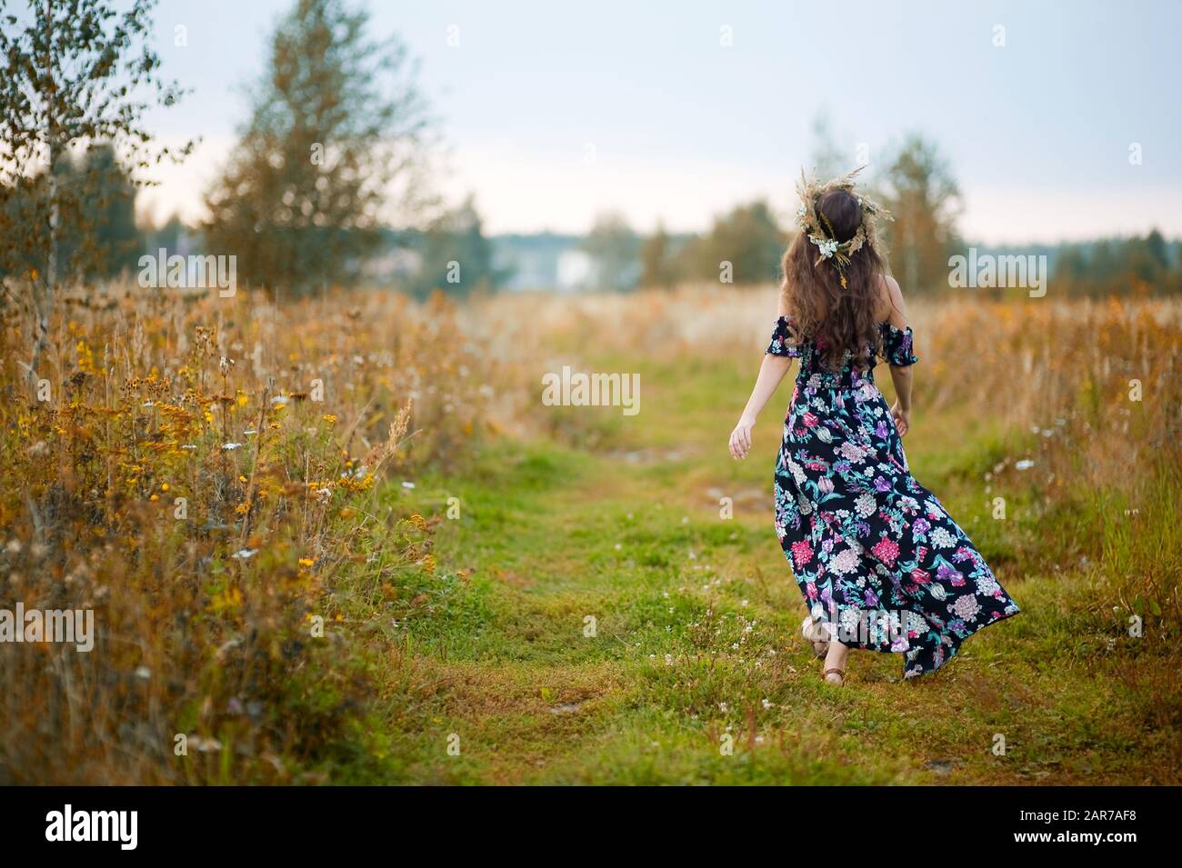 Das junge Mädchen in einer bunten Sonnenkresse mit einem Kranz auf dem Kopf geht auf einer Landstraße weg Stockfoto