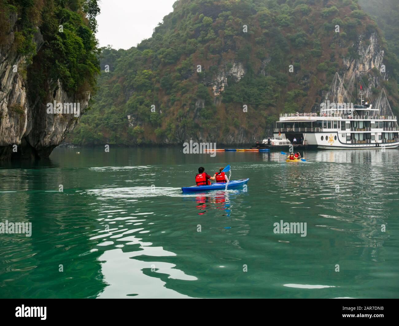 Touristen in Kajaks mit Orchid-Kreuzfahrtschiff und Kalksteinklippen, Halong Bay, Vietnam, Asien Stockfoto