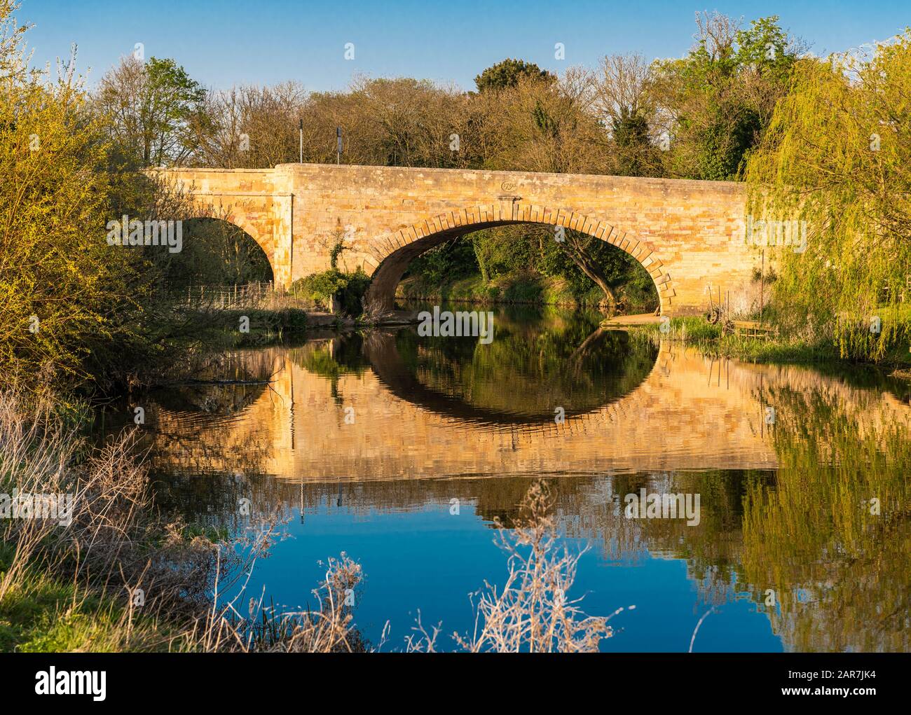 Der Fluss Nene in Wansford, Cambridgeshire, England, Großbritannien, mit Jura-Kalksteinbrücke an einem ruhigen, sonnigen Tag im April und Reflexionen im Wasser Stockfoto