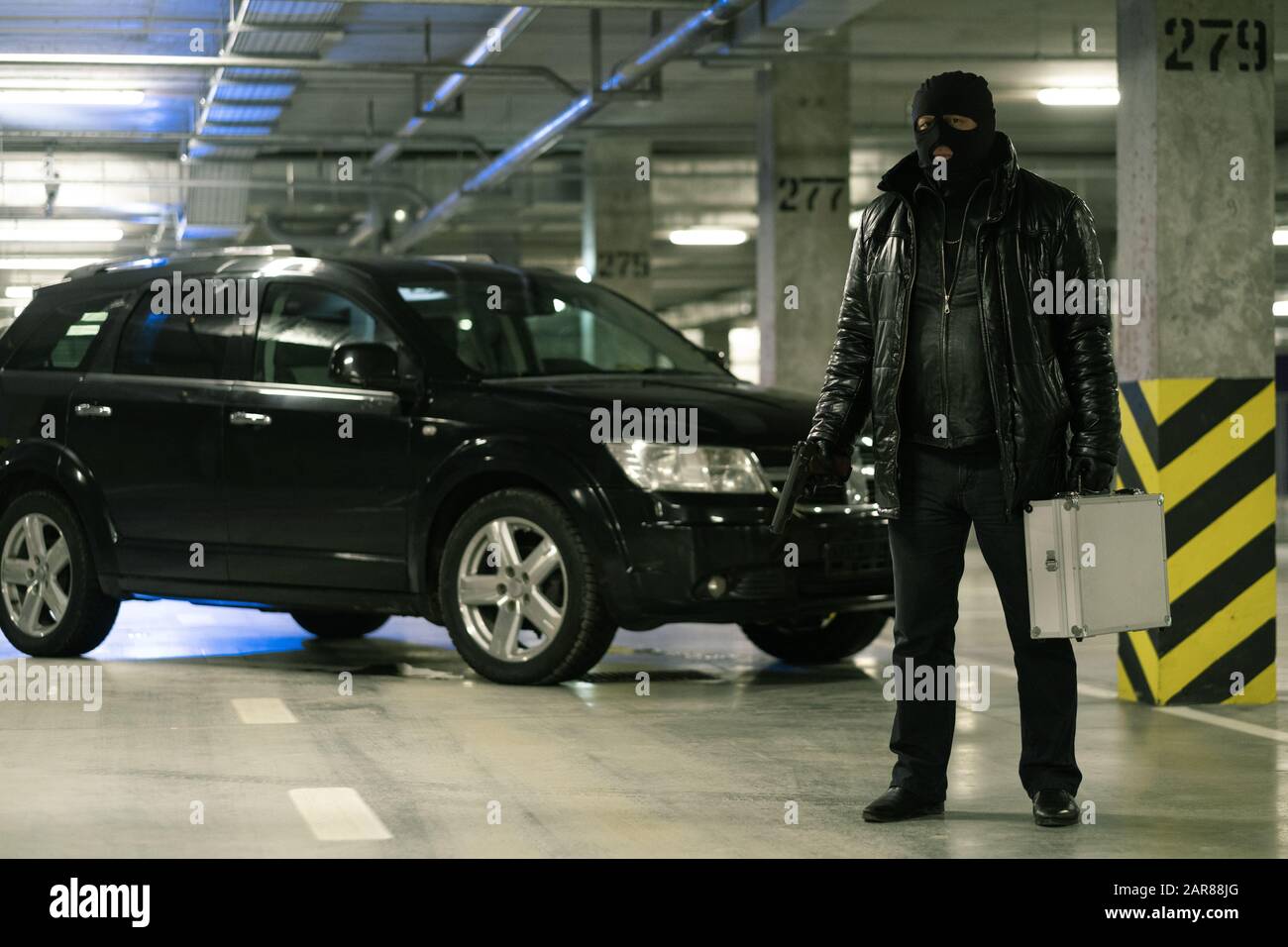 Gangster in schwarzer Jacke und Balaclava auf dem Kopf, der auf dem Hintergrund des Autos steht Stockfoto