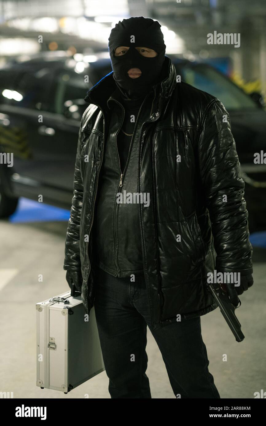 Verbrecher in schwarzer Jacke und Balaclava mit Handfeuerwaffe und Koffer mit Bargeld Stockfoto