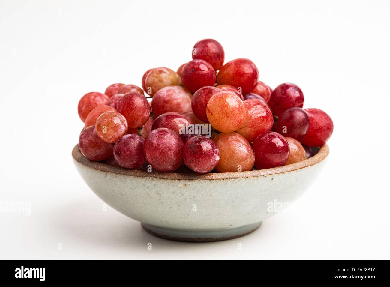 Ein Haufen frischer roter Trauben, die auf einer kleinen Keramikschale auf einem einfarbigen weißen Hintergrund gebündelt sind. Stockfoto