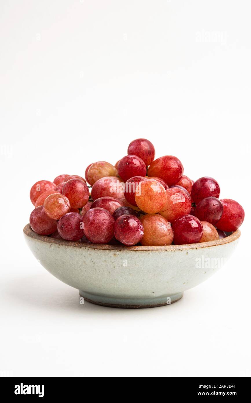 Ein Haufen frischer roter Trauben, die auf einer kleinen Keramikschale auf einem einfarbigen weißen Hintergrund gebündelt sind. Stockfoto