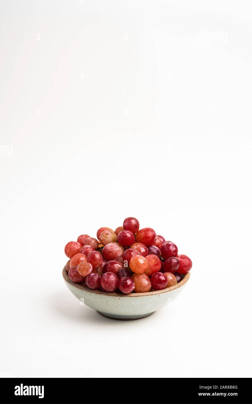 Ein Haufen frischer roter Trauben, die auf einer kleinen Keramikschale auf einem einfarbigen weißen Hintergrund gebündelt sind. Stockfoto