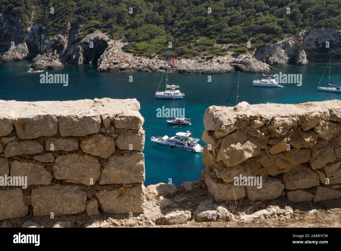 Blick von einer Steinmauer über die Bucht von Cala Llonga mit Booten auf kristallklarem Wasser in Cala Llonga, Ibiza, Balearen, Spanien Stockfoto