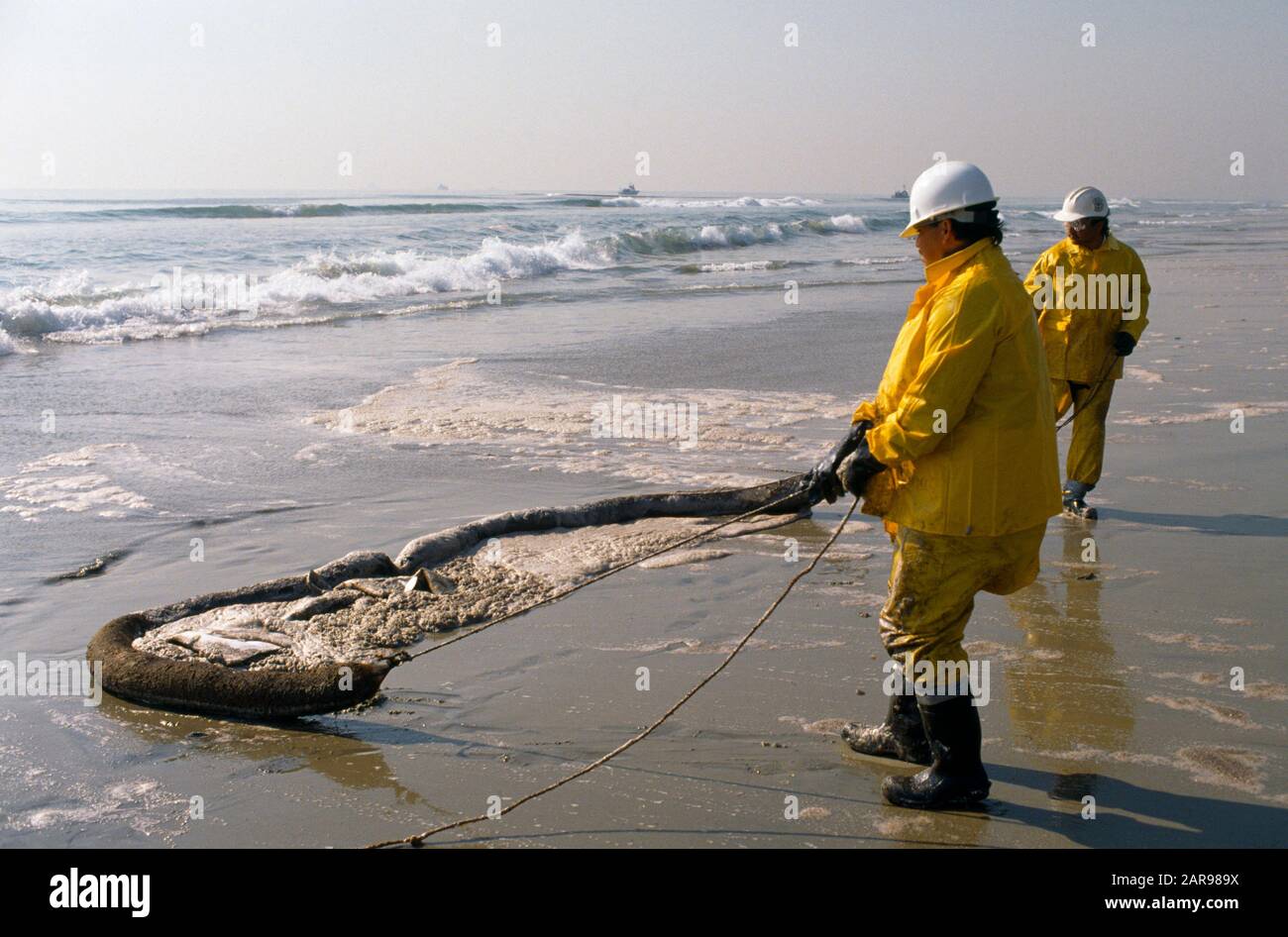 Mit harten Hüten und Sicherheitskleidung ziehen Arbeitsleute an einem handgesteuerten Ausleger, um Ölverschmutzung durch einen Tankerausstoß am Huntington Beach, CA, zu beseitigen. Stockfoto