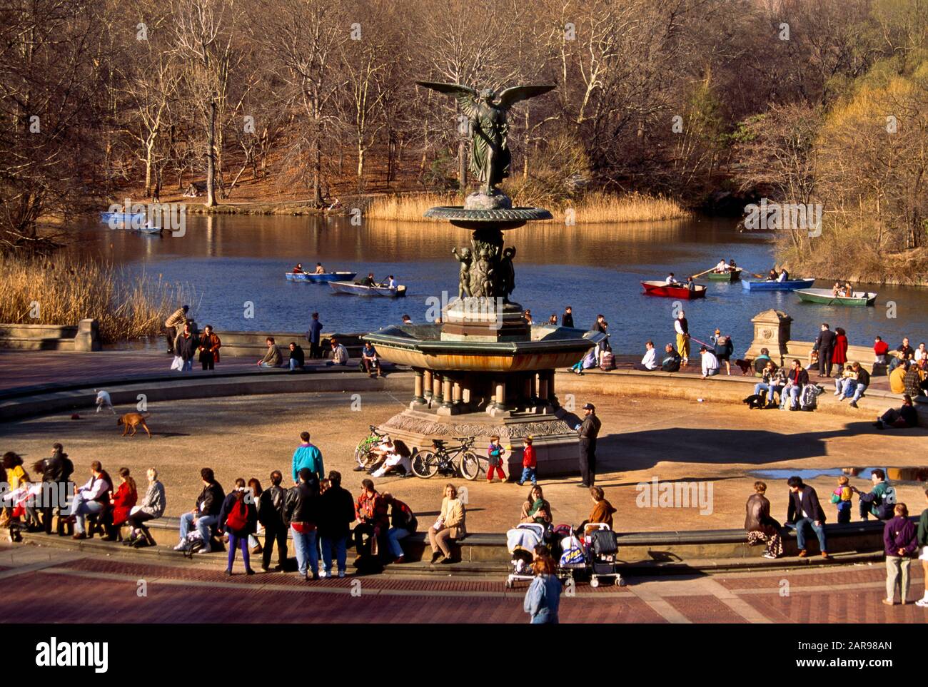 Fußgänger mit mehreren Rassen entspannen sich am Bethesda-Brunnen im Central Park, New York City. Der Brunnen wurde von Emma Stebbins im Jahr 1868 entworfen. Sie war die erste Frau, die eine öffentliche kommission für ein großes Kunstwerk in New York City erhielt. Die bronzene, achtfussige Statue zeigt einen weiblichen geflügelten Engel, der sich auf der Spitze des Brunnens hinaufberührt. Unter ihr befinden sich vier 4-Fuß-Cherubs, die Temperance, Reinheit, Gesundheit und Frieden repräsentieren Stockfoto