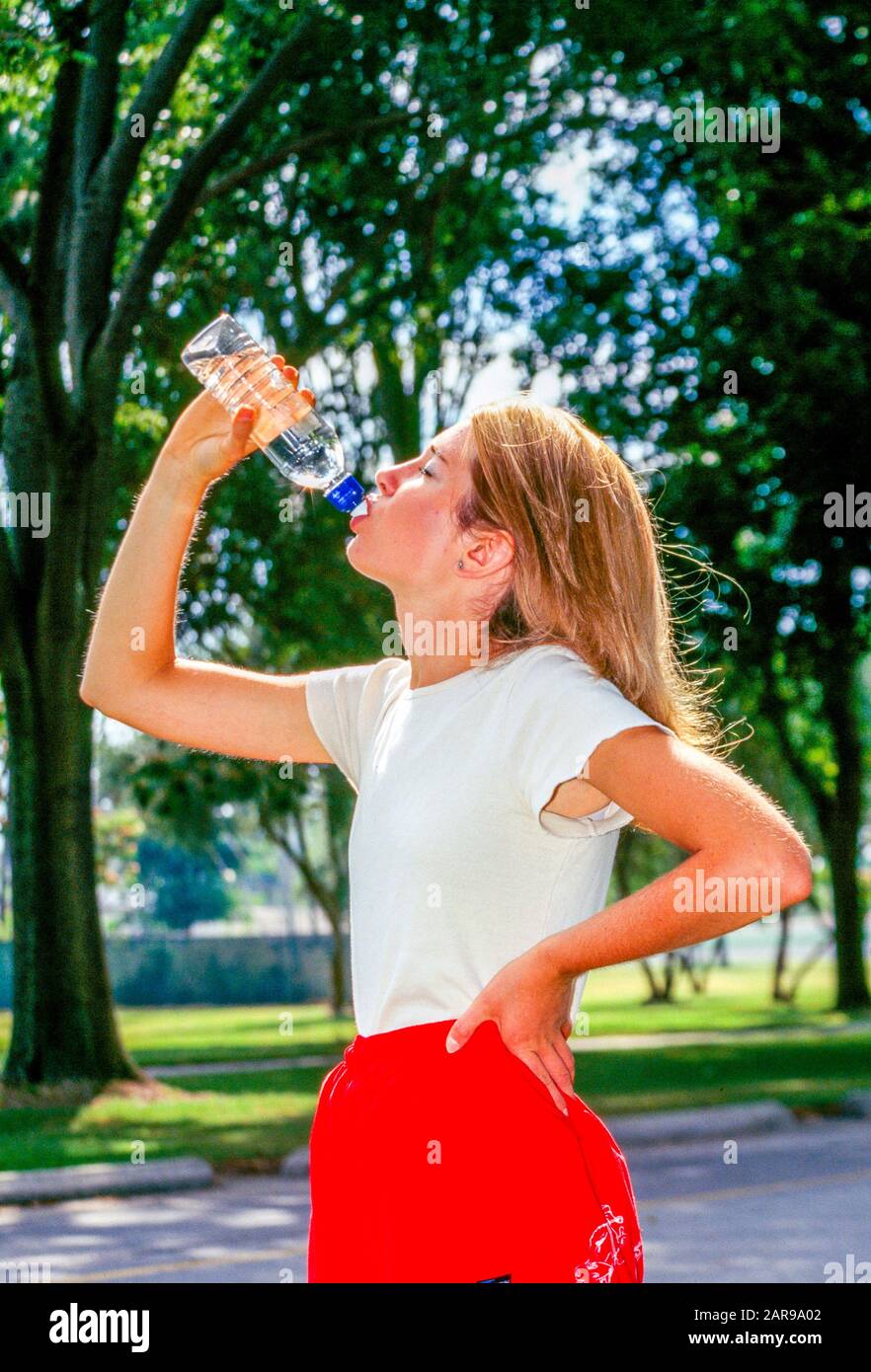Eine blonde junge Erwachsene Joggerin macht eine Pause vom laufen, um ein erfrischendes Getränk aus einer Flasche Wasser in einem Long Beach, CA, City Park MODEL RELEASE zu sich zu nehmen Stockfoto