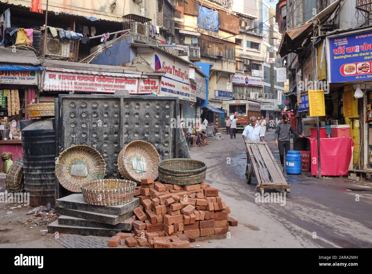 Ein Portier mit einem Kinderwagen, der an einer unordentlichen Szene in Bhuleshwar, Mumbai, Indien, vorbeikommt, eine Baustelle mit Körben von Trägern, die für eine Arbeitspause zur Seite gestellt wurden Stockfoto