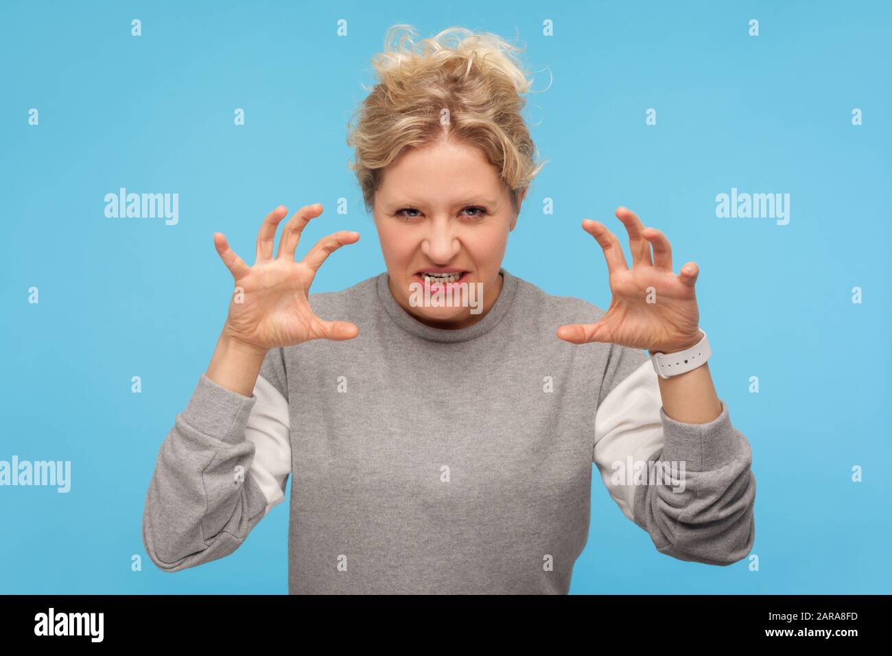 Wütende Frau mit lockigem Haar im Sweatshirt, das mit erhobenen Krallen und geklemmten Zähnen steht und starken Zorn, Reizungen und aggressives e zeigt Stockfoto
