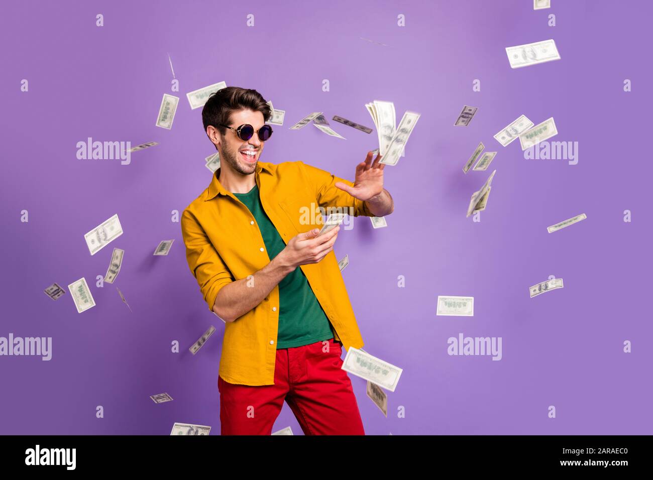 Foto des fröhlichen, aufgeregt weißen trendigen Mannes, der Geld wegwirft und seine wohlhabende lächelnde Zahncreme in roten Hosen zeigt, isoliert violetter Hintergrund Stockfoto