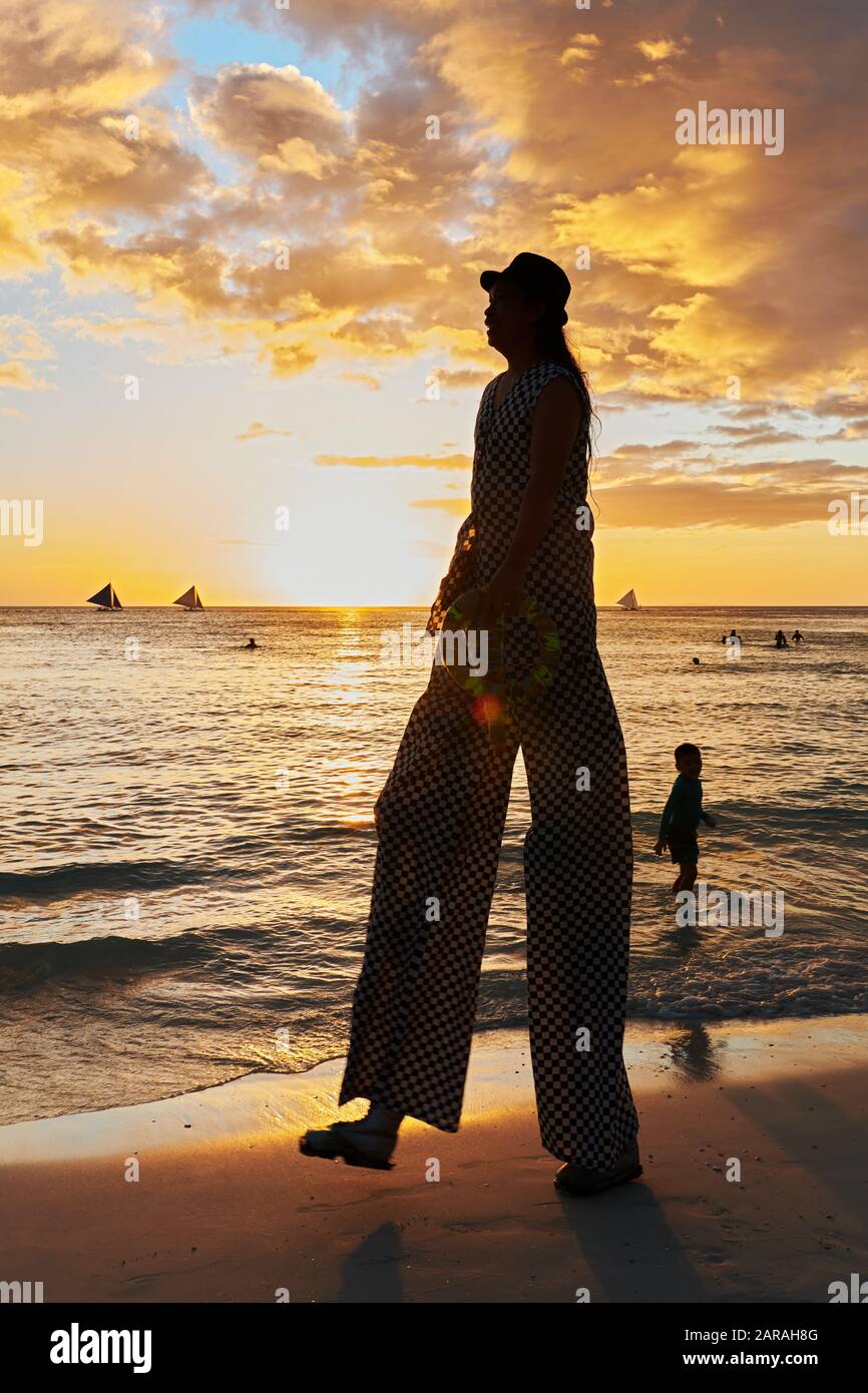 Boracay, Aklan, Philippinen - 1. Januar 2020: Jongleur auf Stelzen, die bei Sonnenuntergang am weißen Strand spazieren gehen, mit einem kleinen Jungen, der ihn beobachtet Stockfoto