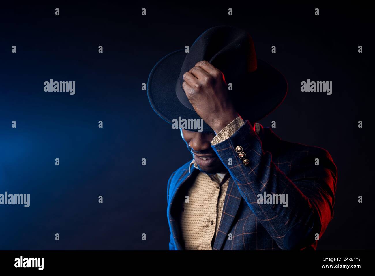 Cooler, dunkelhäutiger Mann in stilvollem Anzug, der seinen schwarzen Hut berührt und nach unten blickt. Nahaufnahme des modischen afroamerikanischen männlichen Modells, das auf dar posiert Stockfoto