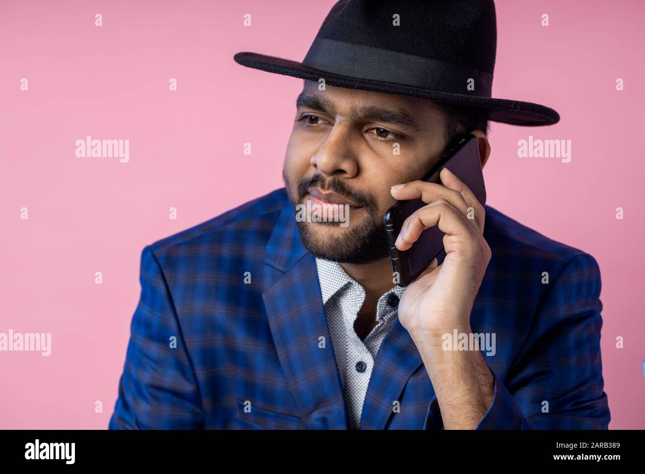 Besorgter junger indischer bärtiger Geschäftsmann, der auf dem Handy spricht. Nahaufnahme des Mannes in formeller Kleidung, schwarzer Hut, mit ernsthaftem, konzentriertem Aussehen während ha Stockfoto