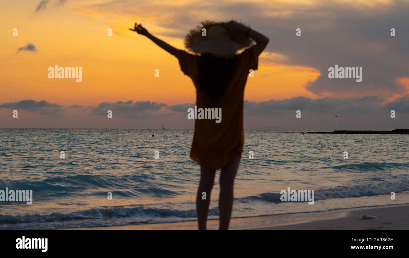 Silhouette der Fit Frau mit langen brünetten Haaren in Strohhut mit erhobenen Armen, die bei Sonnenuntergang an der Küste des Ozeans jubeln. Stockfoto