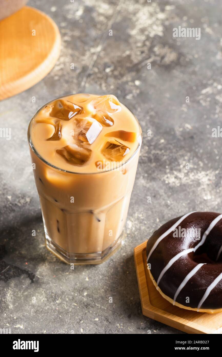 Eiskaffee mit Eiswürfeln in einem hohen Glas und einem frischen Schokoladendonut. Erfrischendes Getränk auf dem grauen Hintergrund des Tisches. Nahaufnahme des selektiven Fokus. Stockfoto