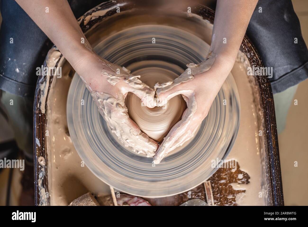 Ein Potter arbeitet in der Werkstatt mit Ton auf einem Potter-Rad. Frauenhände zeigen ein Herzschild. Das Konzept der Liebe zur Töpferkunst und Kreativität. Oben Stockfoto