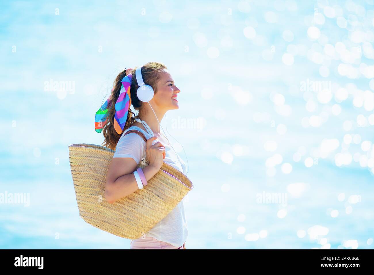 Lächelnde Frau im mittleren Alter in weißem T-Shirt und pinkfarbenen Shorts mit Strandabschnitt Musik mit Kopfhörern an der Seeküste hören. Stockfoto