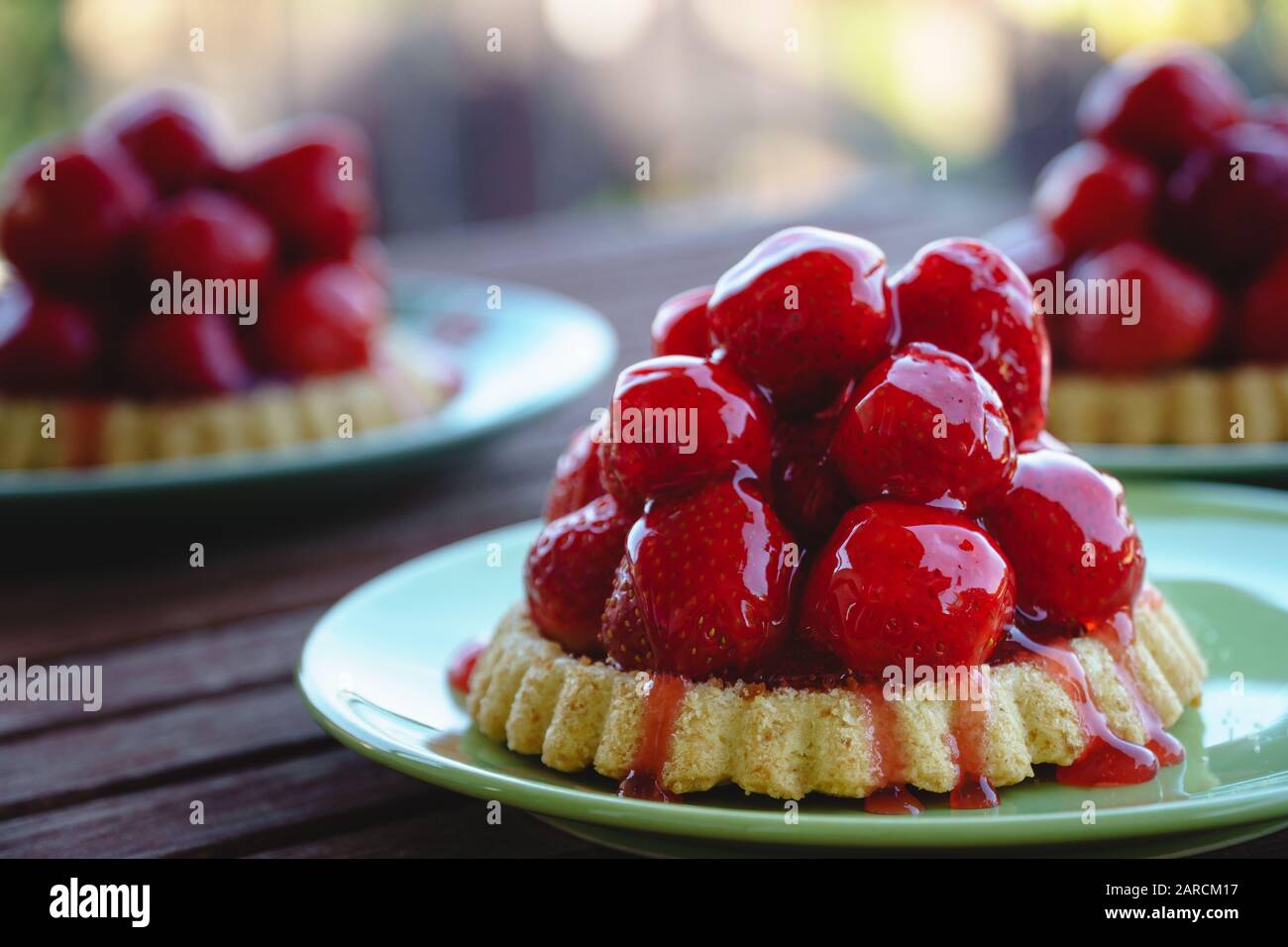 Drei frische vegane Erdbeertarts, serviert auf grünen Platten auf einem Holztisch auf dem Balkon - Seitenansicht, horizontale Ausrichtung, verschwommener Hintergrund Stockfoto