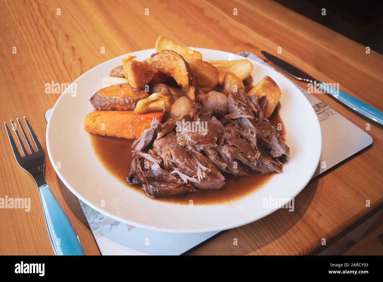 Hausgemachtes, langsam gebratenes Lamm mit Wurzelgemüse und Gravy auf einem weißen Teller auf einem Holztisch in einer Küche. Stockfoto
