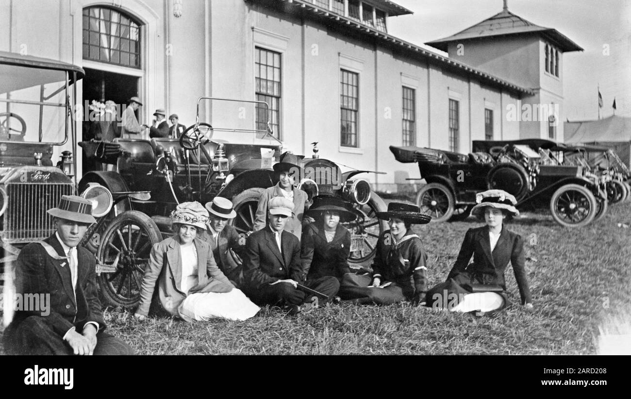 1900ER 1910ER-JAHRE-GRUPPE MÄNNER FRAUEN, DIE IM GRAS SITZEN, MIT GEPARKTEN AUTOS HINTER TRIBÜNEN DES ÖRTLICHEN FAIRGROUND-ZIRKUSZELTES IM HINTERGRUND - M8932 HAR001 HARS KOPIE SPACE FRIENDSHIP HALBE LÄNGE DAMEN AUTO MÄNNER UNTERHALTUNG TRANSPORT B&W GEPARKTES AUGE KONTAKT WEITWINKEL 8 GLÜCK FREIZEIT STILE AUTOS FREIZEIT STOLZ AUF LOKALE FAIRGROUNDS KONZEPTIONELLE TRIBÜNEN AUTOMOBILE STYLISCHE FAHRZEUGE ACHT MODE ENTSPANNUNG ZUSAMMENGEHÖRIGKEIT SCHWARZ-WEISS KAUKASISCHE ETHNIE HAR001 ALTMODISCH Stockfoto