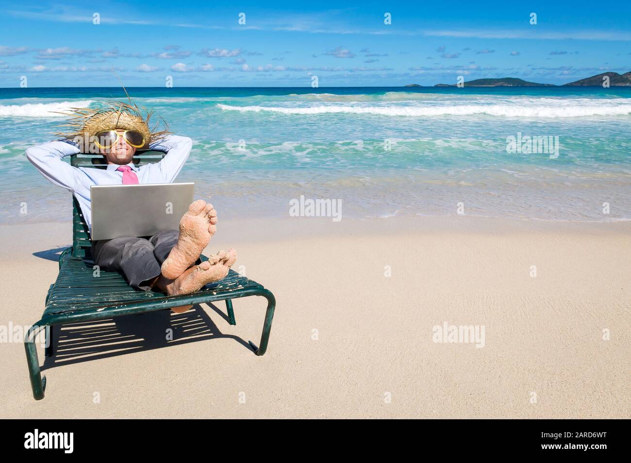 Barfuß-Geschäftsmann tritt mit Laptop auf dem Schoß und Sonnenbrille und Strohhut auf den Kopf zurück in einen Strandstuhl Stockfoto