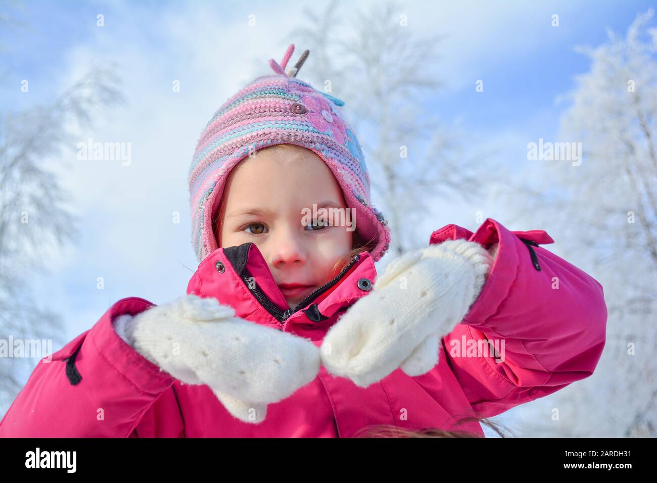Porträt des jungen Mädchens in Winterkleidung und buntem Wollhut, mit den Händen in weißen Wollhandschuhen, gegen blauen Himmel und weiße, gefrorene TR Stockfoto