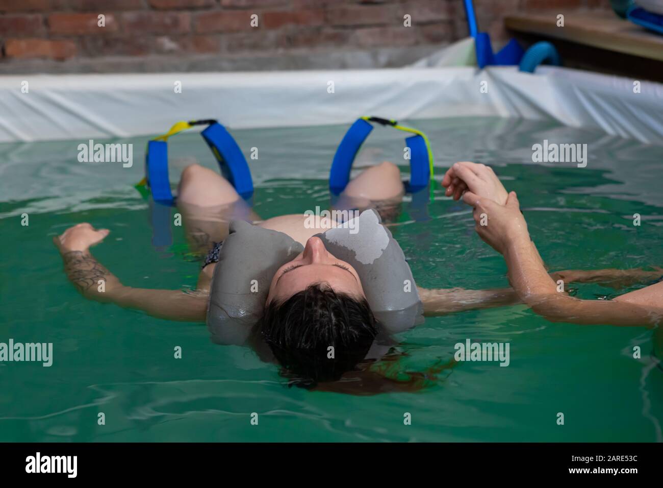 Junge schwangere Frau erholt sich im Pool mit Gummihalskissen und Beinauflage schwimmt beim Training im kleinen Schwimmbad Stockfoto