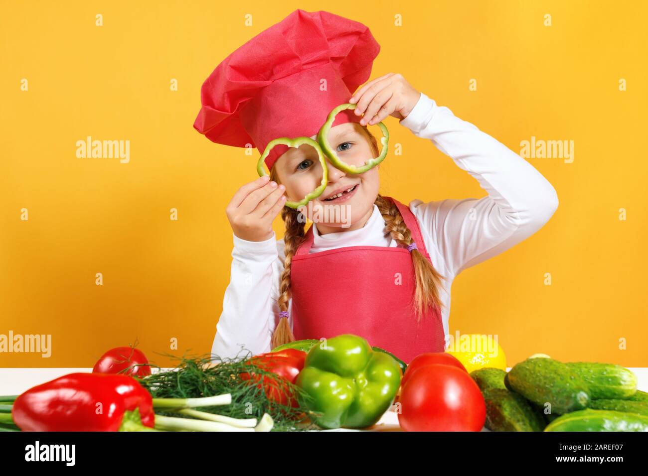 Ein fröhliches Kind in der Mütze eines Kochs hat Spaß mit Gemüse und hält Pfeffer vor seinen Augen, als wäre es in einer Brille. Kleines Mädchen sitzt an einem Tisch auf einem Bac Stockfoto