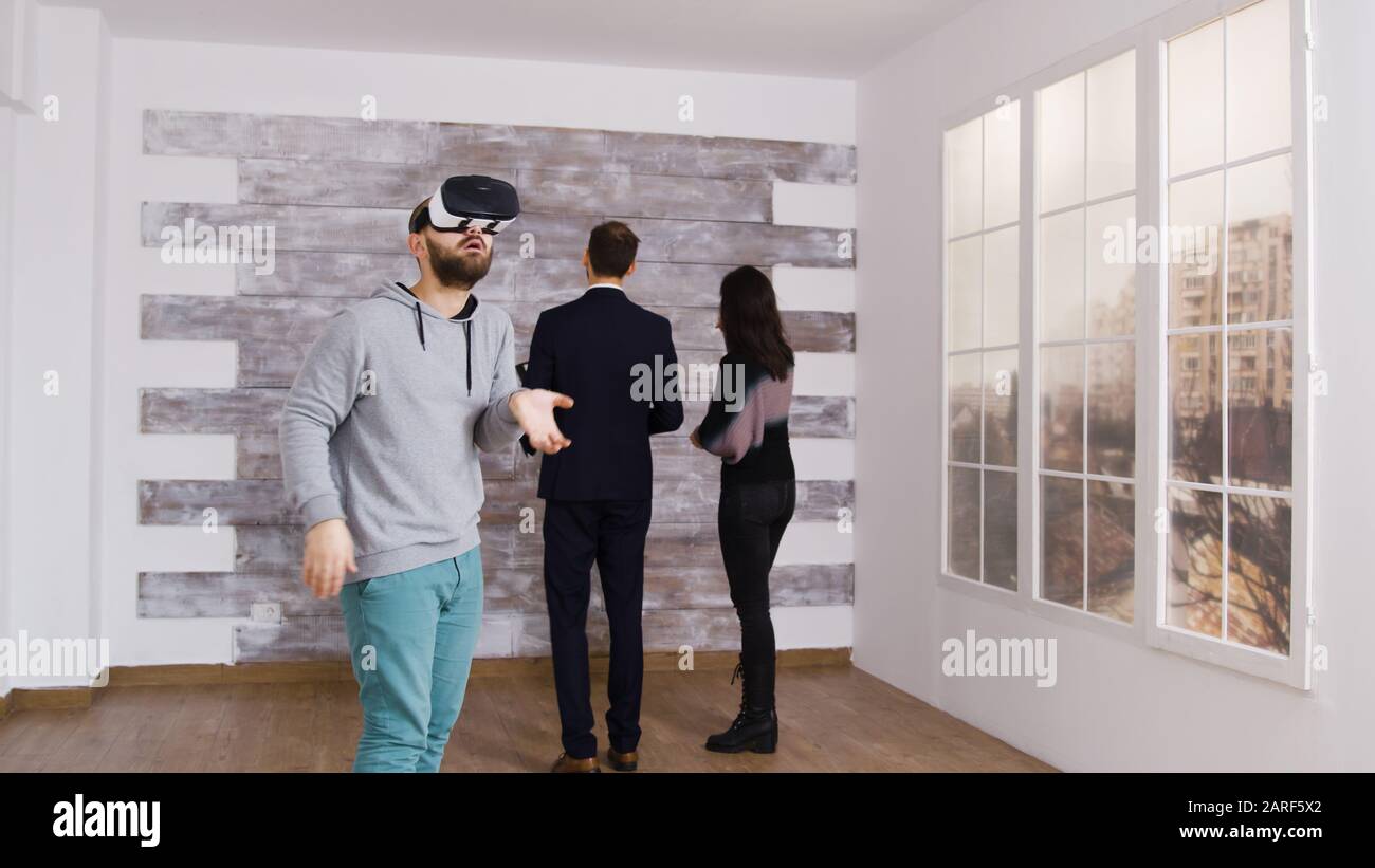 Kaukasischer Mann mit Virtual-Reality-Headset in leerer Wohnung, während eine Frau mit Immobilienmakler spricht. Stockfoto