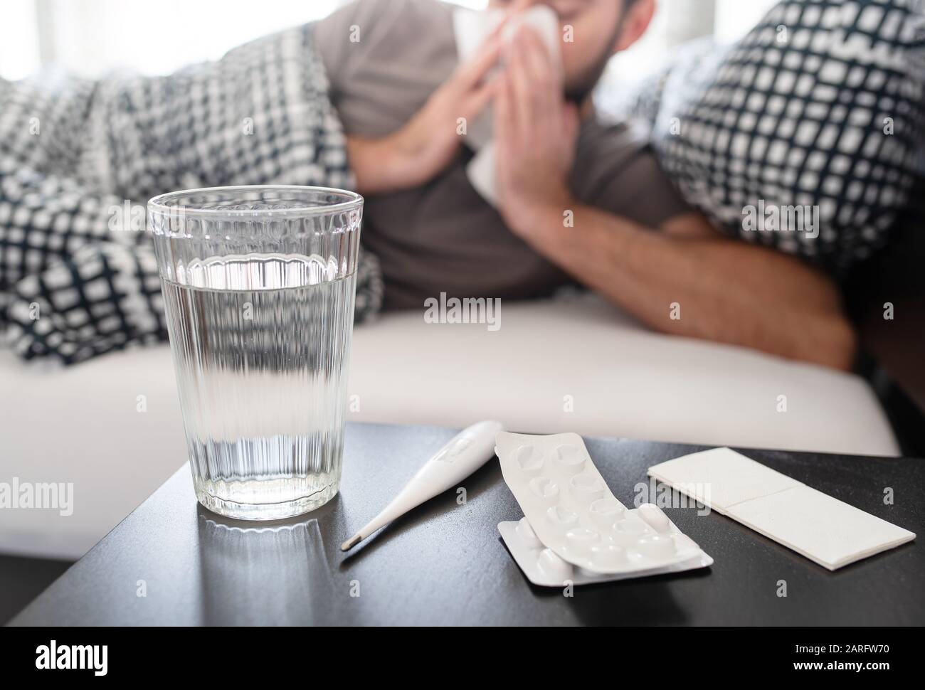 Medikamente, Thermometer und Glas Wasser auf Nachttisch gegen kranke männliche Person, die im Bett liegt und an Erkältung oder Grippeid leidet und seine Nase bläst Stockfoto