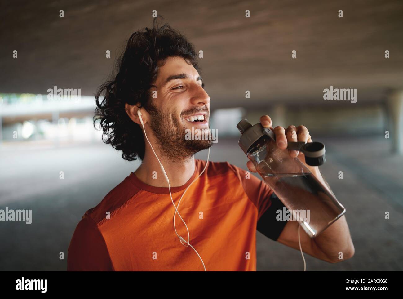 Porträt eines lächelnden fröhlichen jungen Mannes mit Ohrhörern Trinkwasser aus wiederverwendbarer Flasche im Freien in der Stadt Stockfoto