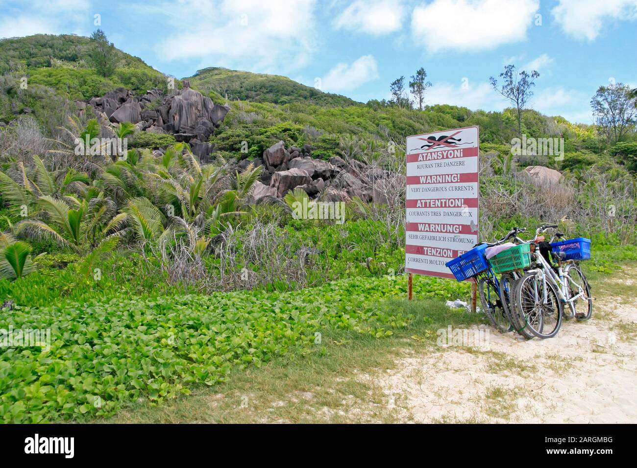 Fahrräder, die vor warnungsgefährlichen Strömungen geparkt wurden, signieren mit Wasserburschen und Palmen auf der Insel La Digue auf den Seychellen. Stockfoto