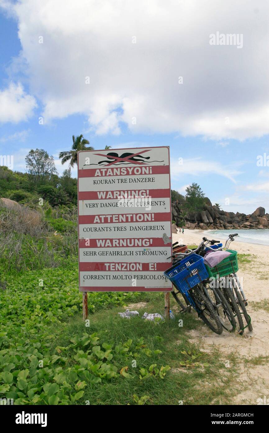 Fahrräder, die vor warnungsgefährlichen Strömungen geparkt wurden, signieren mit Wasserburschen und Palmen auf der Insel La Digue auf den Seychellen. Stockfoto