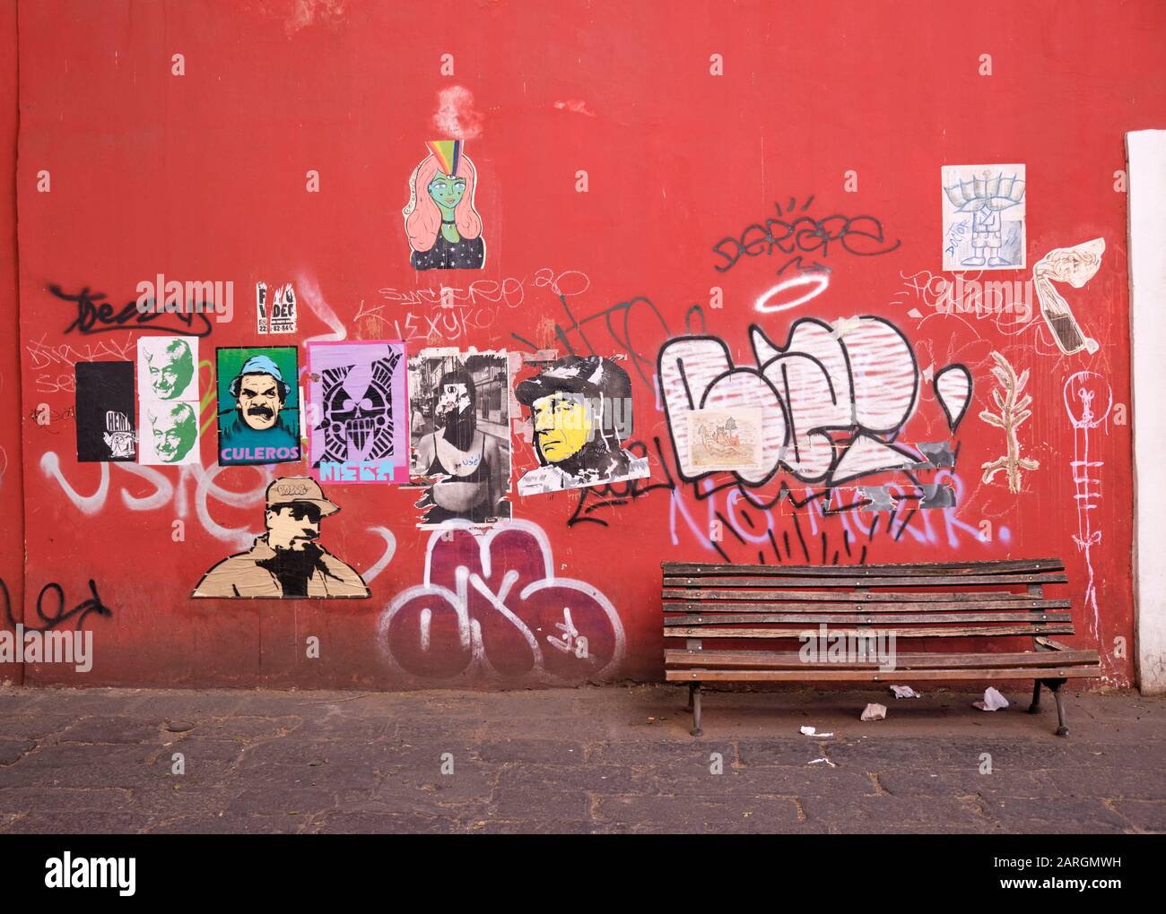 Rote Wand in der kolonialen Altstadt von Puebla mit verschiedenen Dekal-Kunstplakaten und Graffiti mit leerer Holzbank bedeckt Stockfoto