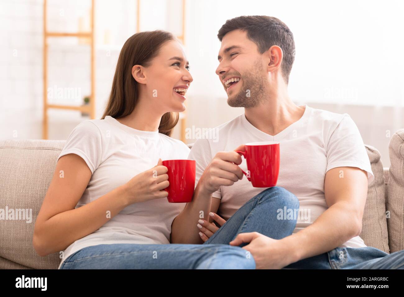 Ein Paar Lacht, Wenn Man Heiße Getränke Auf Der Couch Zu Hause Sitzt Stockfoto