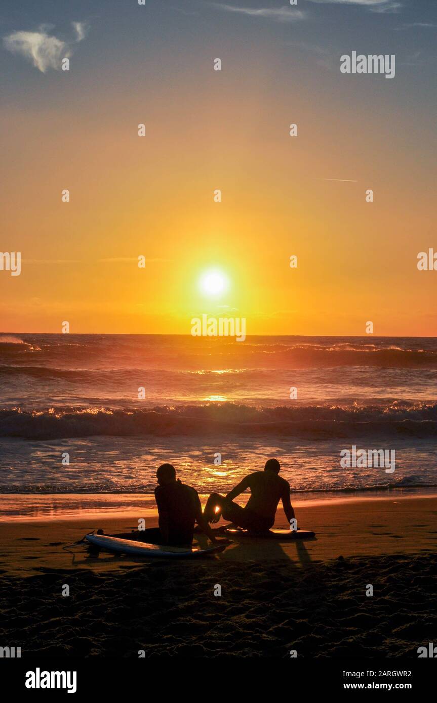 Silhouetten von zwei Surfern, die bei Sonnenuntergang am Strand sitzen Stockfoto