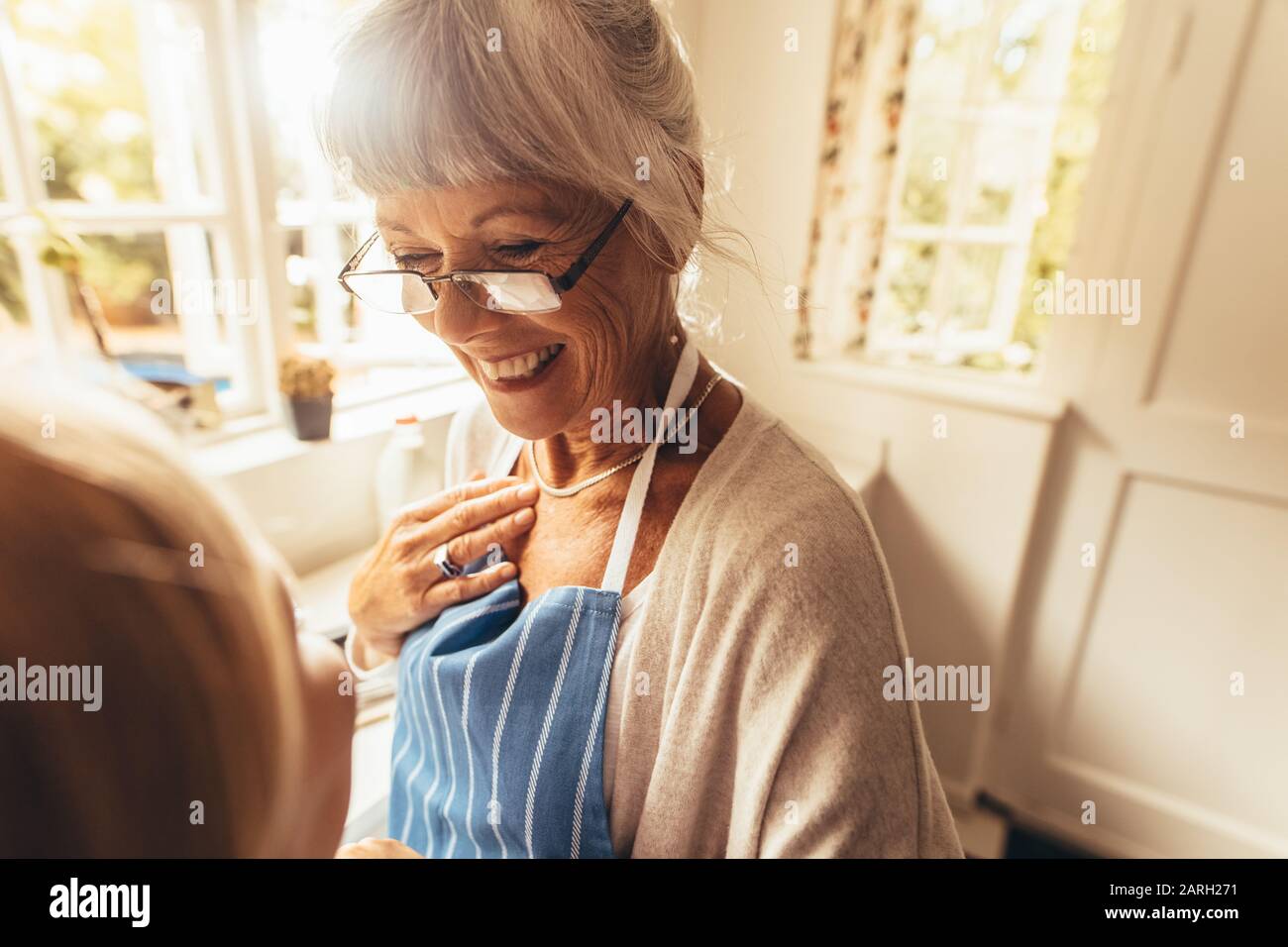 Lächelnde Seniorin, die in ihrem Haus in einer Schürze steht. Fröhliche alte Frau, die in der Küche steht und lächelt. Stockfoto