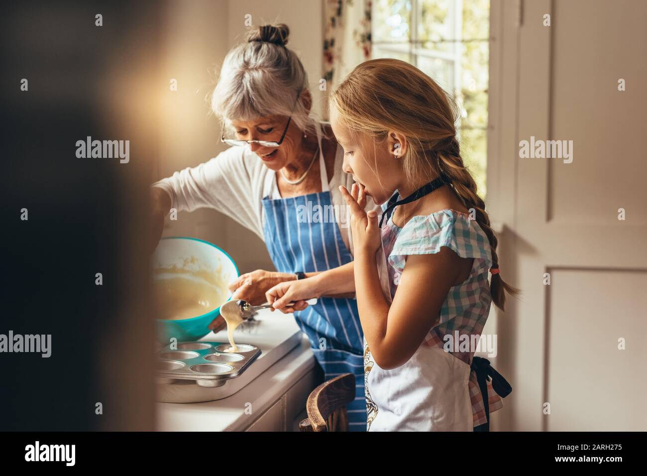 Großmutter unterrichte dem Kind, Tassenkuchen zu machen. Fröhliche Großmutter und Kind gießen Kuchenbatter in Tassenkuchformen. Stockfoto