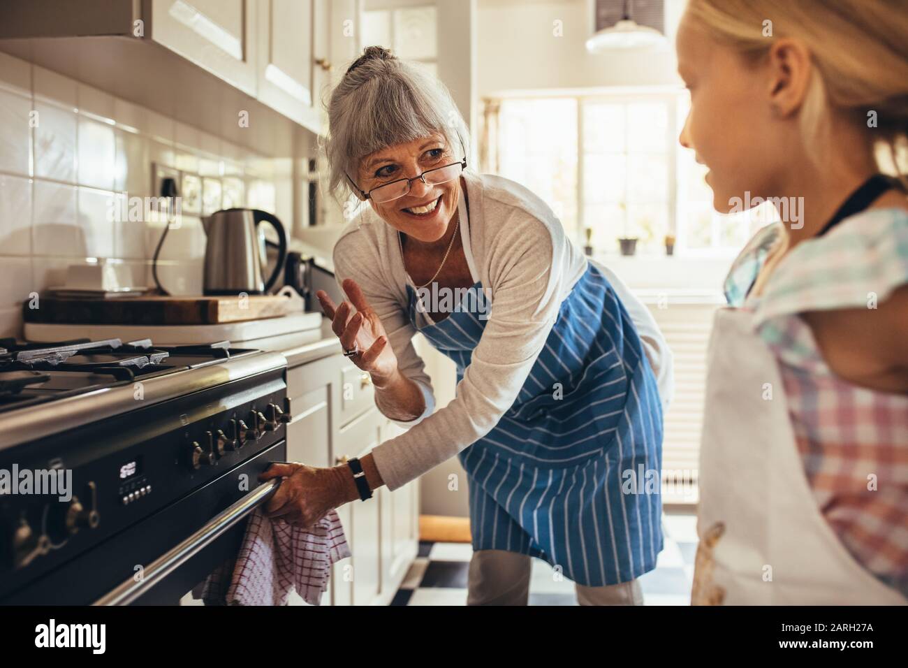 Lächelnde Seniorin in der Schürze, die die Backofentür öffnet. Fröhliche Granny und Kind in der Küche zusammen kochen. Stockfoto