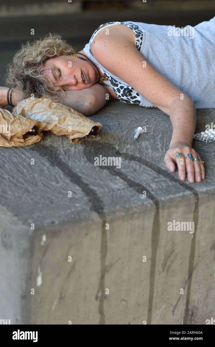 Eine betrunkene, obdachlose Frau, die unter einer Brücke mit Flaschen Alkohol und anderen Drogen um sie herum schläft Stockfoto