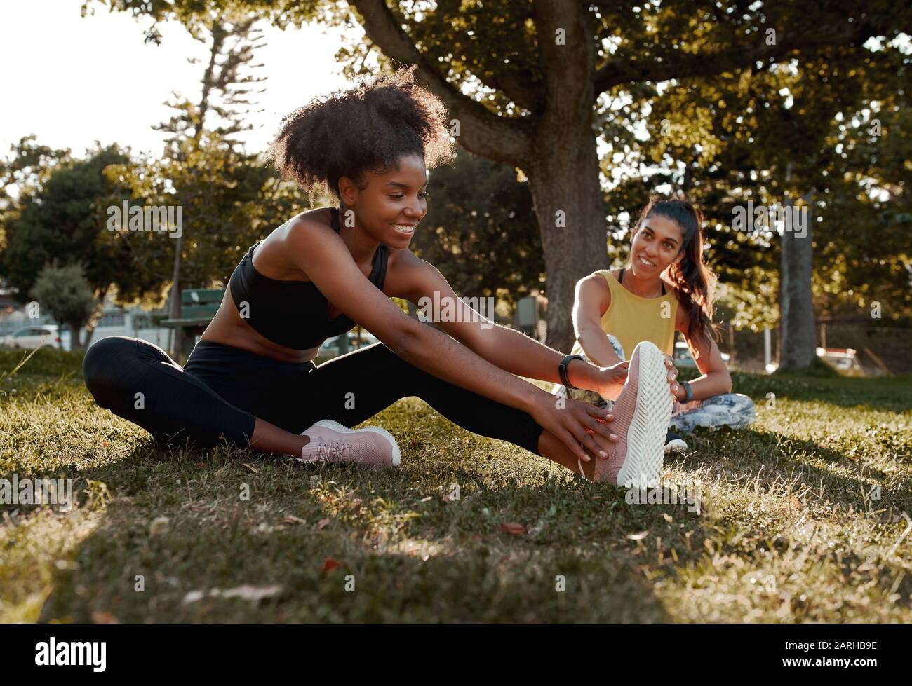 Lächelnd zwei diverse sportliche fit junge weibliche Freunde, die sich im Park die Beine Strecken - afroamerikanische schwarze Frauen erwärmen sich mit ihrem Freund Stockfoto