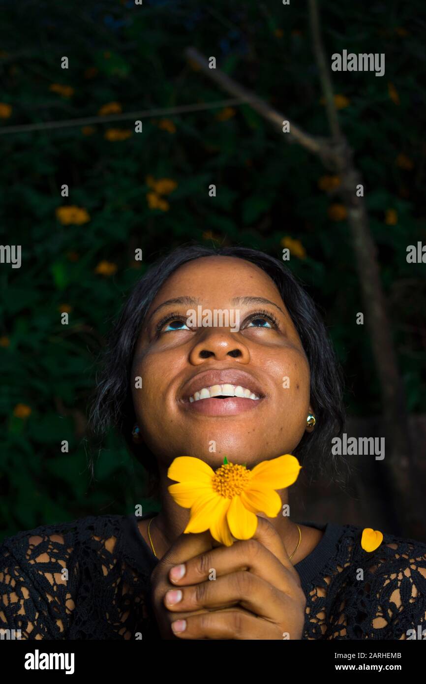 Junge schwarze Frau, die eine Blume mit einigen gefallenen Kronblättern hält, sie liebt mich, sie liebt mich nicht, schaut nach oben und lächelt Stockfoto
