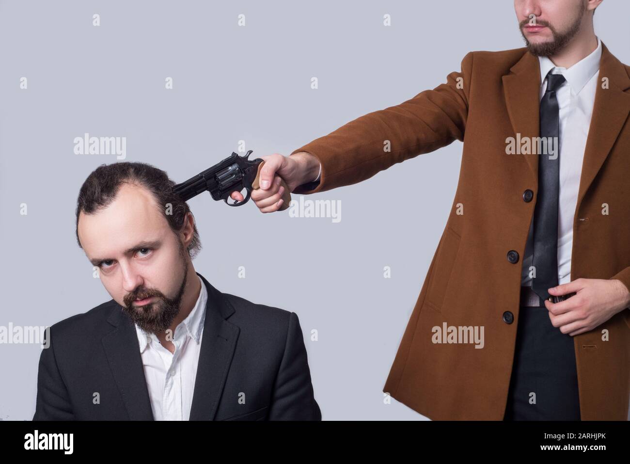 Business man in einem Anzug, der eine Waffe auf den Kopf eines anderen Mannes in einem weißen Hemd zielt, auf grauem Hintergrund. Das Konzept des Tötens. Geschäftliche Tötung. Crimi Stockfoto