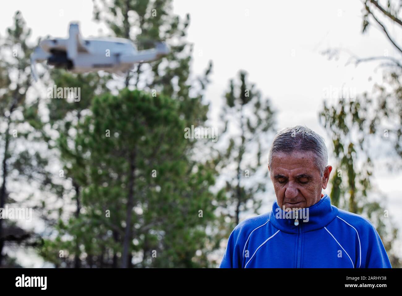 Die Seitenansicht des alten Mannes, der die Drohne mit Blick auf die Fernbedienung fliegt, während er im Park gegen die Bäume steht, konzentriert sich auf den alten Mann Stockfoto