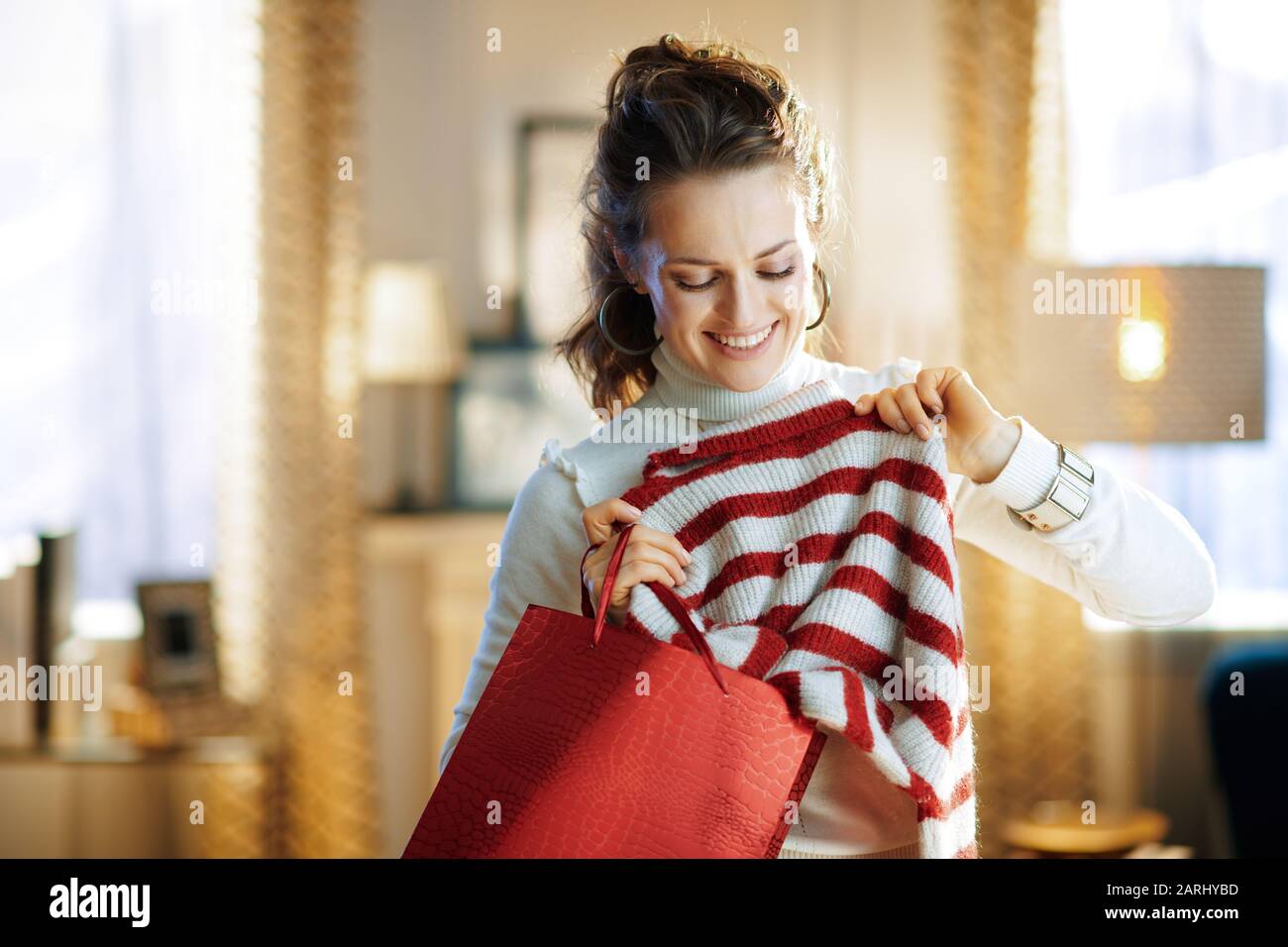 Lächelnde Hausfrau im mittleren Alter in weißem Pullover und Rock mit roter Einkaufstasche, die den Pullover im modernen Zuhause im sonnigen Wintertag gekauft hat. Stockfoto